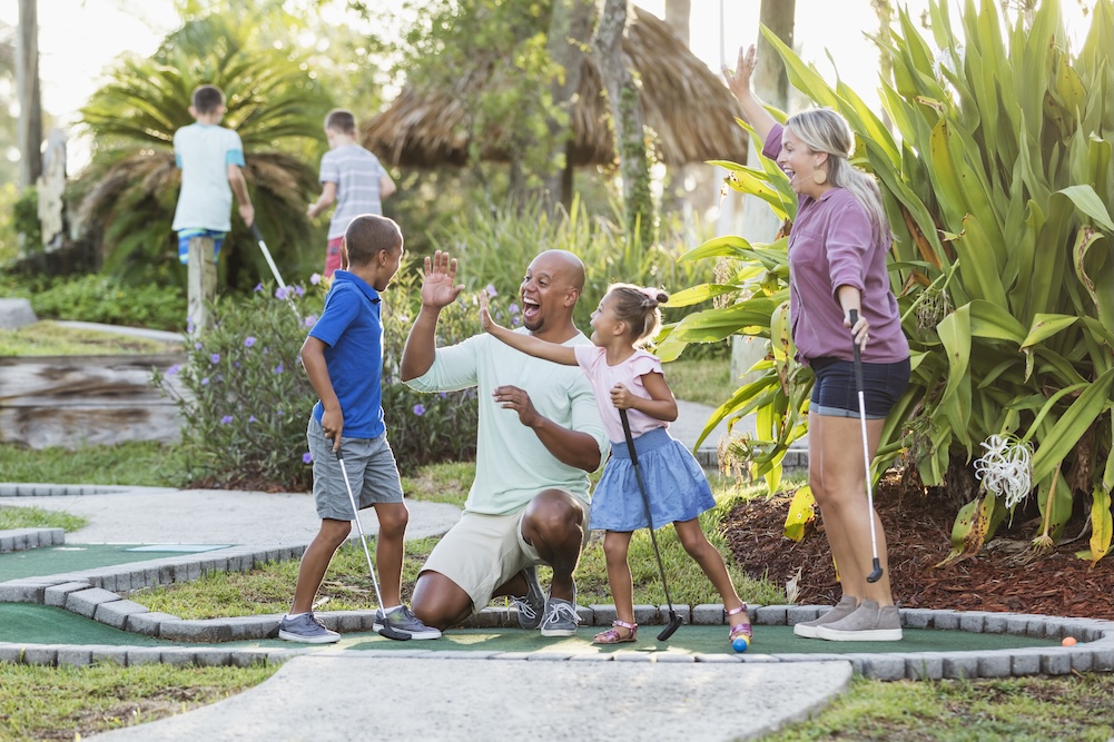 An interracial family having fun together playing miniature golf. The African-American father and Caucasian mother are in their 30s. The children are 9 and 5 years old. Dad and sister are giving the boy high fives for his winning putt.