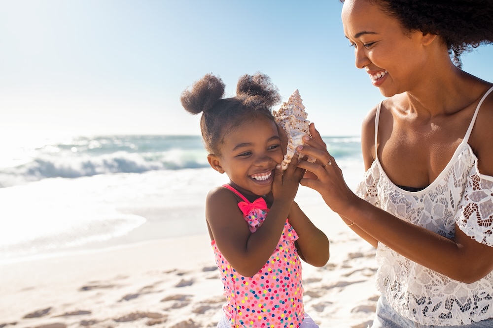 little girl listening to shell on beach with mom