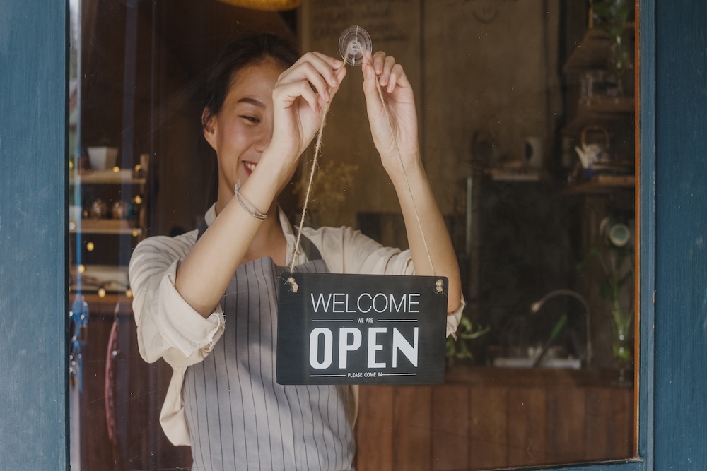 Young Asia manager girl changing a sign from closed to open sign on door cafe looking outside waiting for clients after lockdown. Owner small business, food and drink, business reopen again concept.